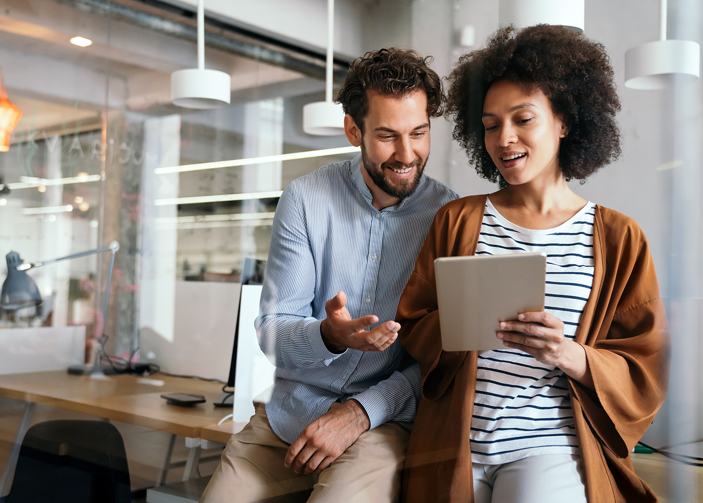 Eine Frau und ein Mann sitzen auf einer Tischkante und besprechen lächelnd einen unbekannten Inhalt auf einem Tablet in ihrer Hand. 